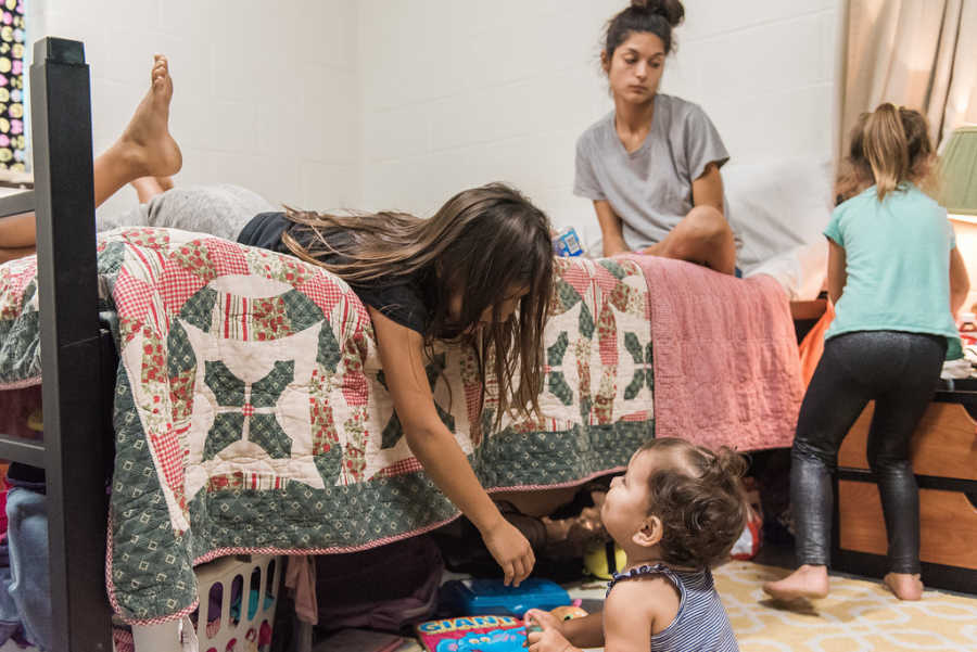 Mother sits on bed with daughter who is leaning over the bed looking at baby sister in homeless shelter