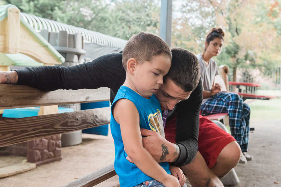 Father sits on bench hugging son at playground of homeless shelter