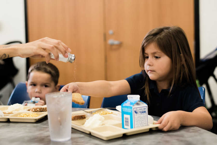 Toddler sits at table eating at homeless shelter while someone salts her food