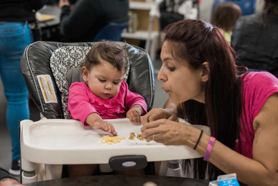 Mother crouches next to daughter in high chair blowing on her food in homeless shelter