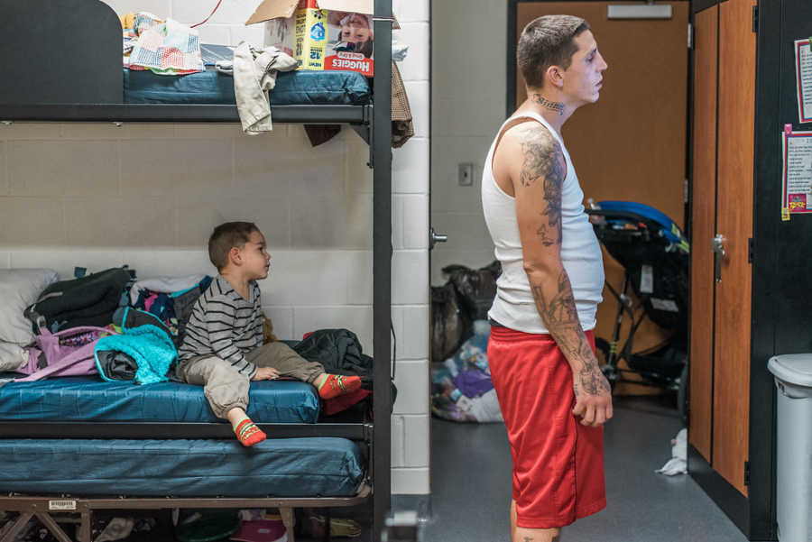 Father stands watching something while son sits behind him on bottom bunk in homeless shelter