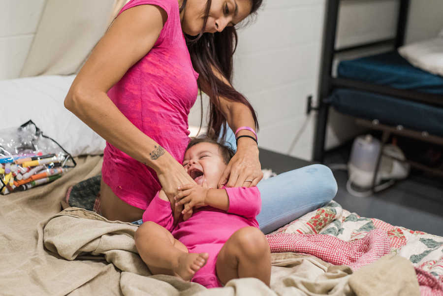 Mother sits on bed tickling infant daughter while she laugh in homeless shelter