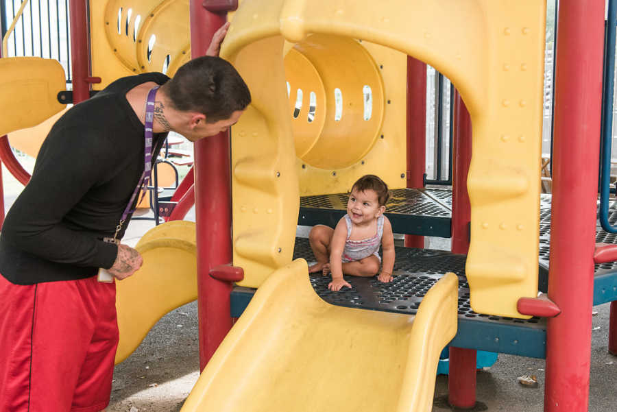 Father smiles at toddler child who is sitting on play structure smiling at homeless shelter