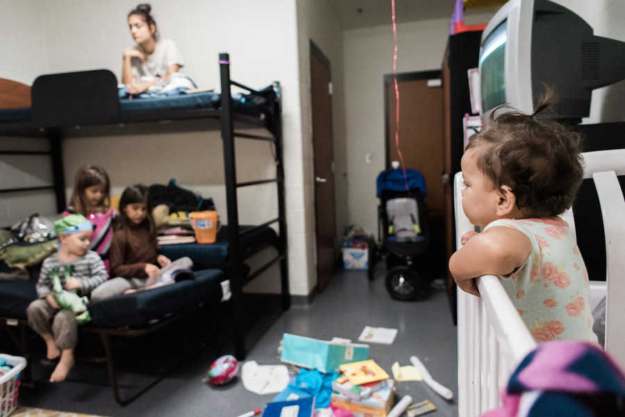 View from corner of homeless shelter room with baby in crib, three kids on bottom bunk, and mother on top bunk