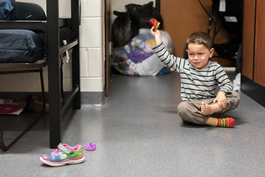 Young boy sits on ground crying while he puts on sock in homeless shelter room 