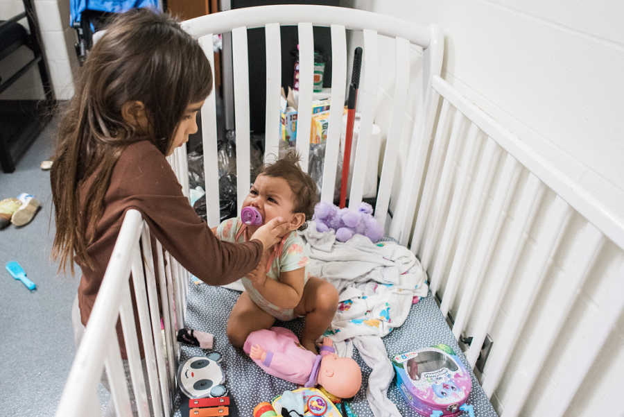 Baby sits in crib with toys while older sister leans over touching her cheek in homeless shelter