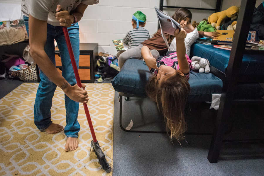 Mother sweeps floor of homeless shelter while three kids lay on bottom bunk of bed