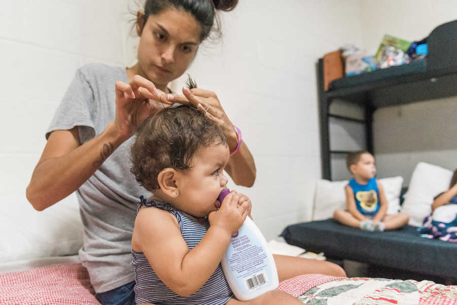 Mother sits doing infant's hair while she bites a bottle in homeless shelter room