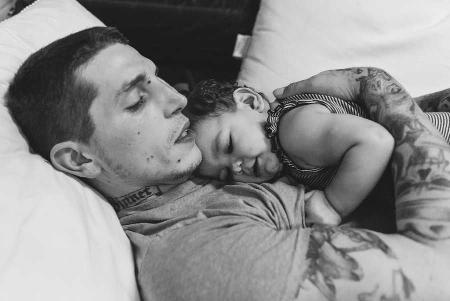 Father lays down in bed holding baby daughter to chest in homeless shelter