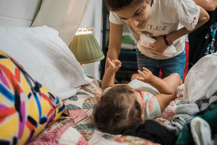 Mother looks over baby daughter lying in bed about to change her diaper in homeless shelter