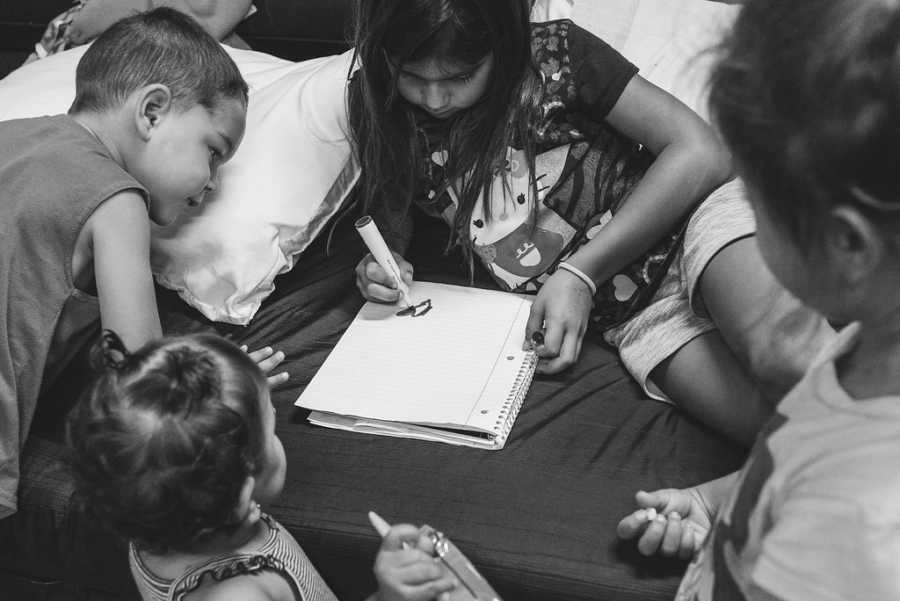 Older sister draws on notebook while three younger siblings watch in homeless shelter