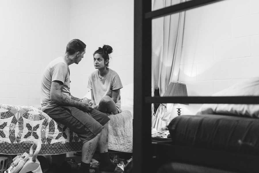 Husband and wife sit on bed of homeless shelter looking at each other