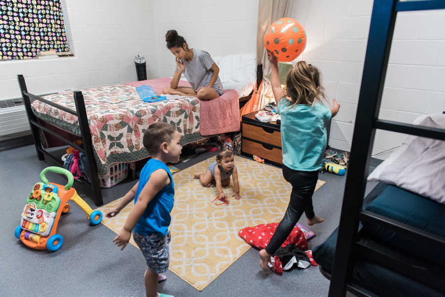 Mother sits on bed while three children play next to her in room in homeless shelter