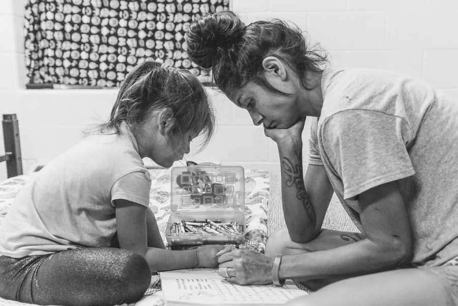 Mother and daughter sit on bed coloring in homeless shelter room