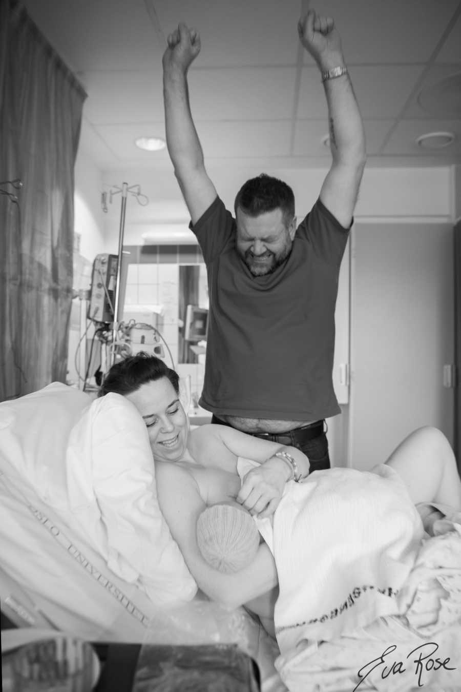 Husband stands with arms in air beside wife who is in hospital bed breastfeeding