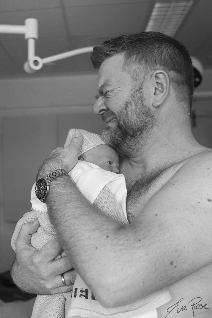 Shirtless father holds newborn to chest crying