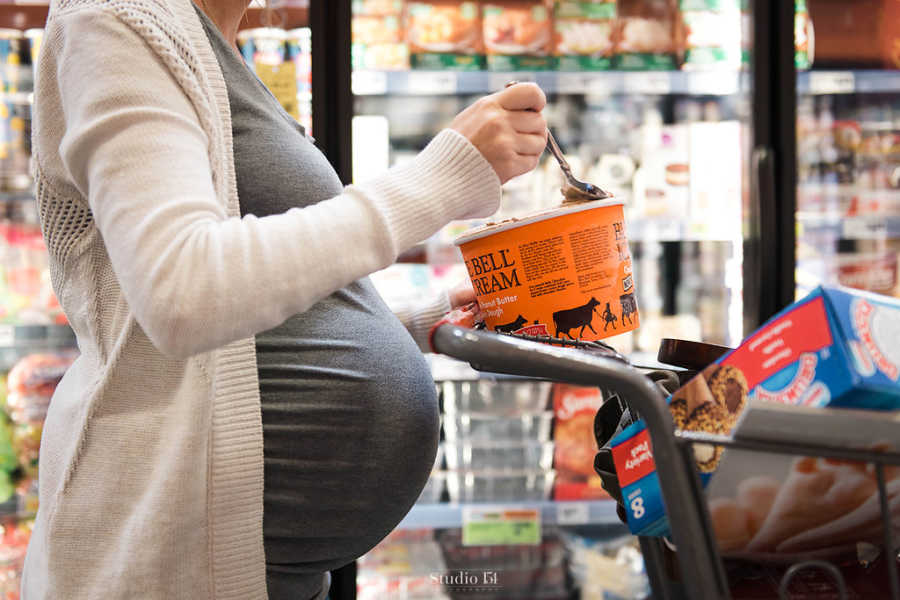 Side view of pregnant woman pushing shopping cart while scooping out bite of ice cream from carton