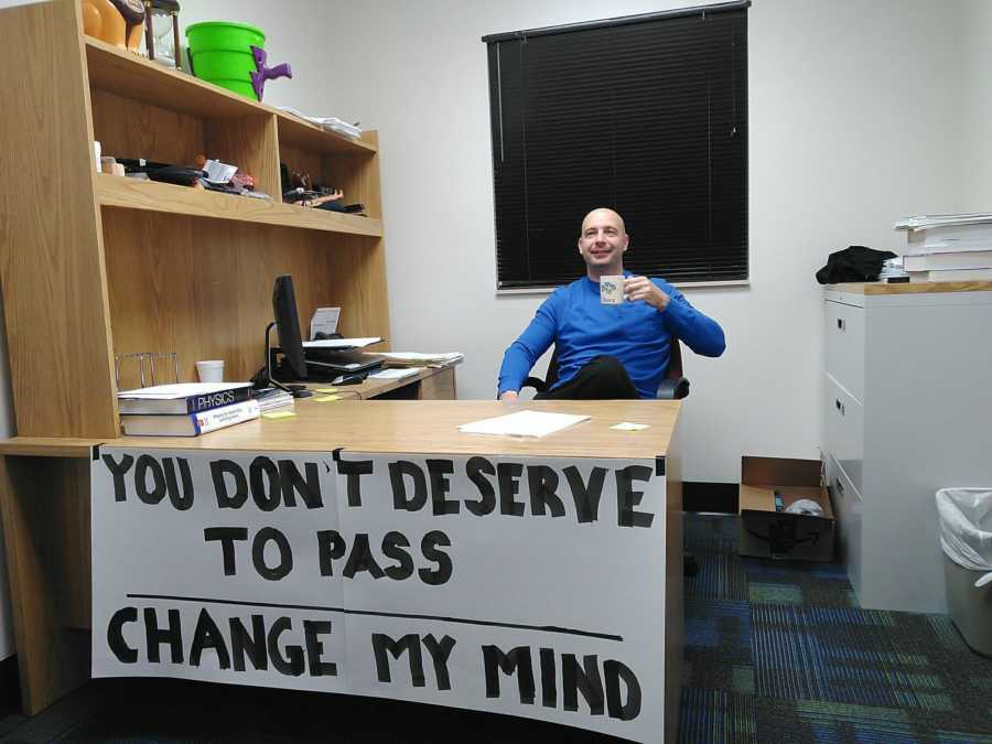 Teacher sitting at desk with sign attached to it saying, "You don't deserve to pass change my mind"