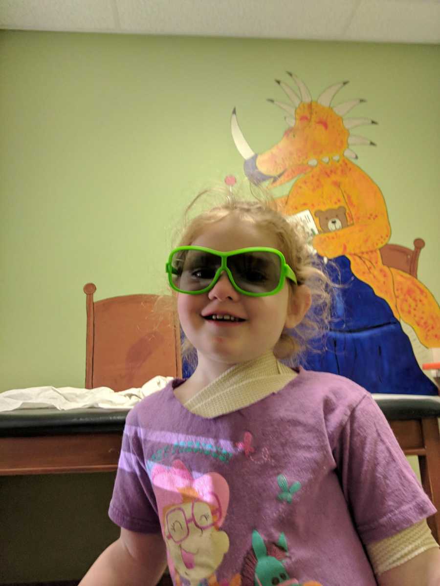 Toddler sitting in doctor's waiting room with sunglasses on and bandages on her body from skin graft