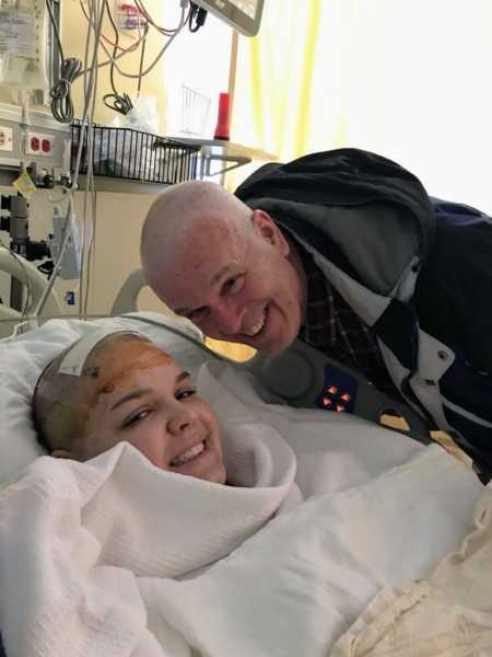 21 year old who had brain surgery lays in hospital bed with father leaning over her who shaved his head for her