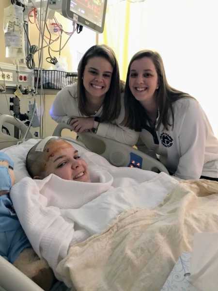 21 year old who had brain surgery lies in hospital bed with sisters standing over her smiling