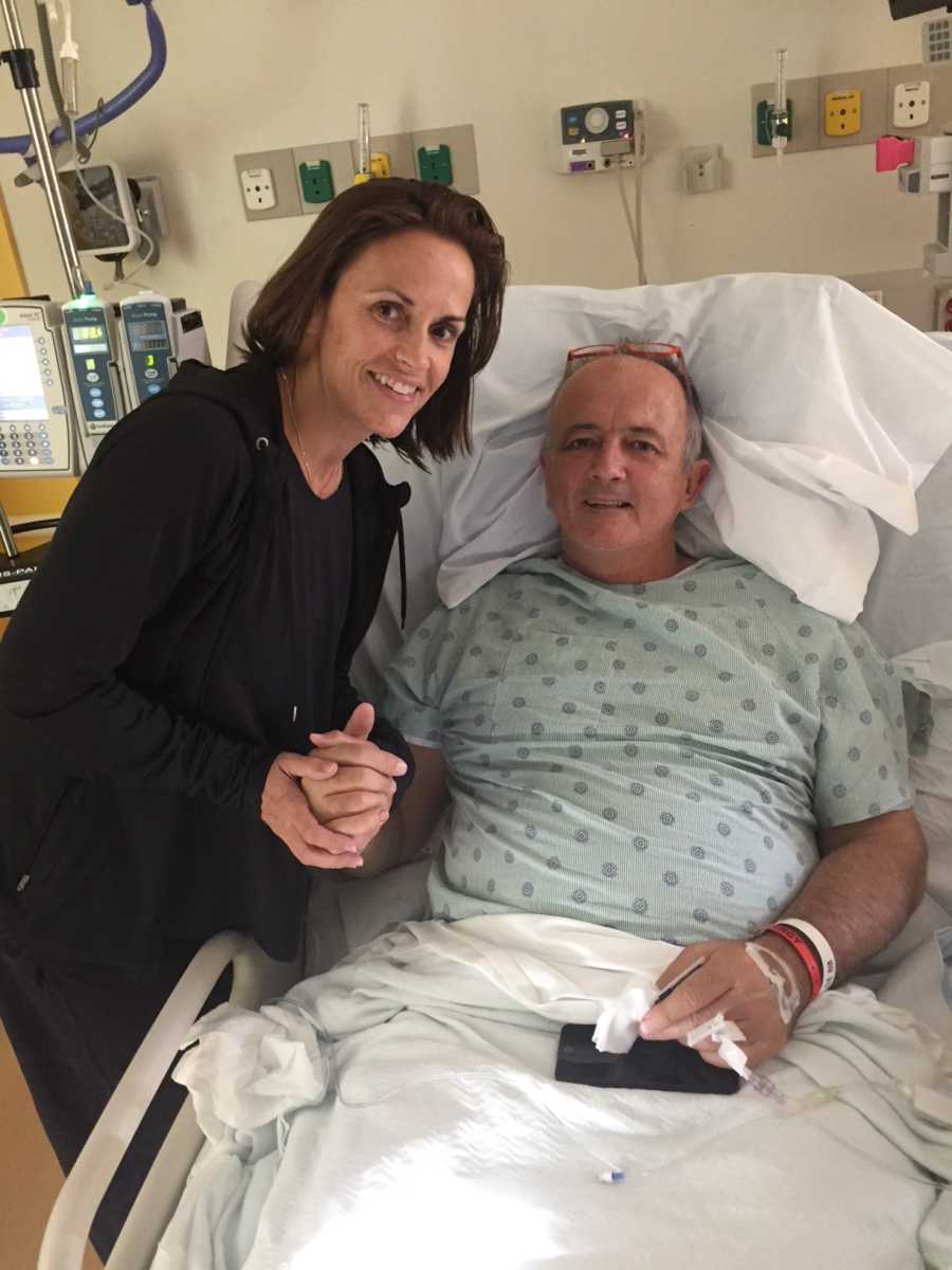 Man whose new kidney came from wife's friend, lays in hospital bed smiling and holding donor's hand