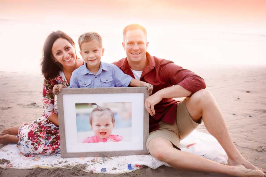 Mother, father, and son sit on beach holding picture frame of daughter they lost to Neuroblastoma Cancer