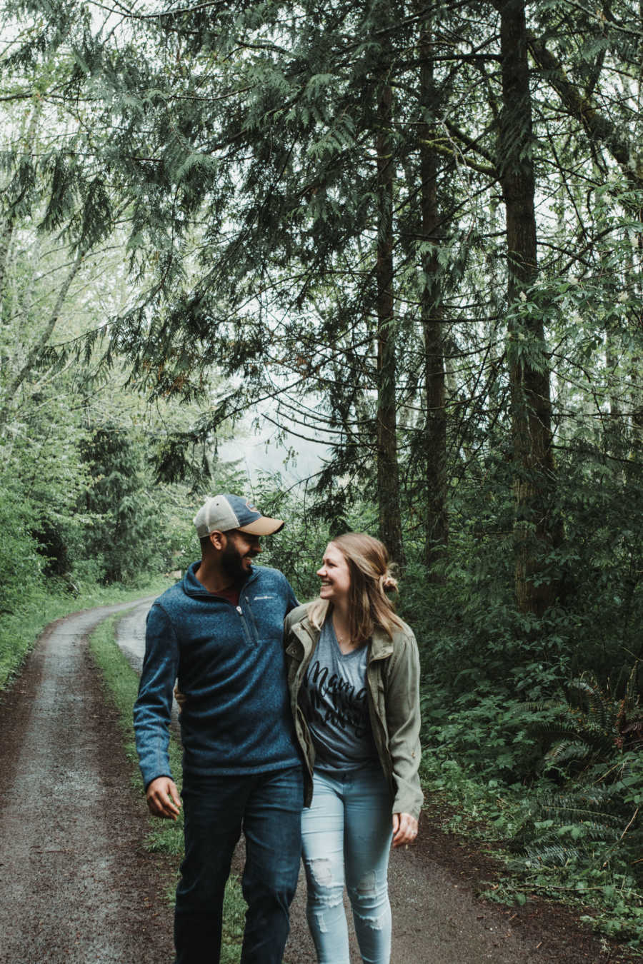 Soon to be adoptive parents smile at each other arm in arm as they walk on a path in forest