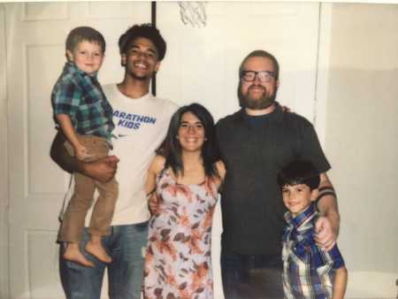 Young man stands next to foster parents and their young son while he holds their other son