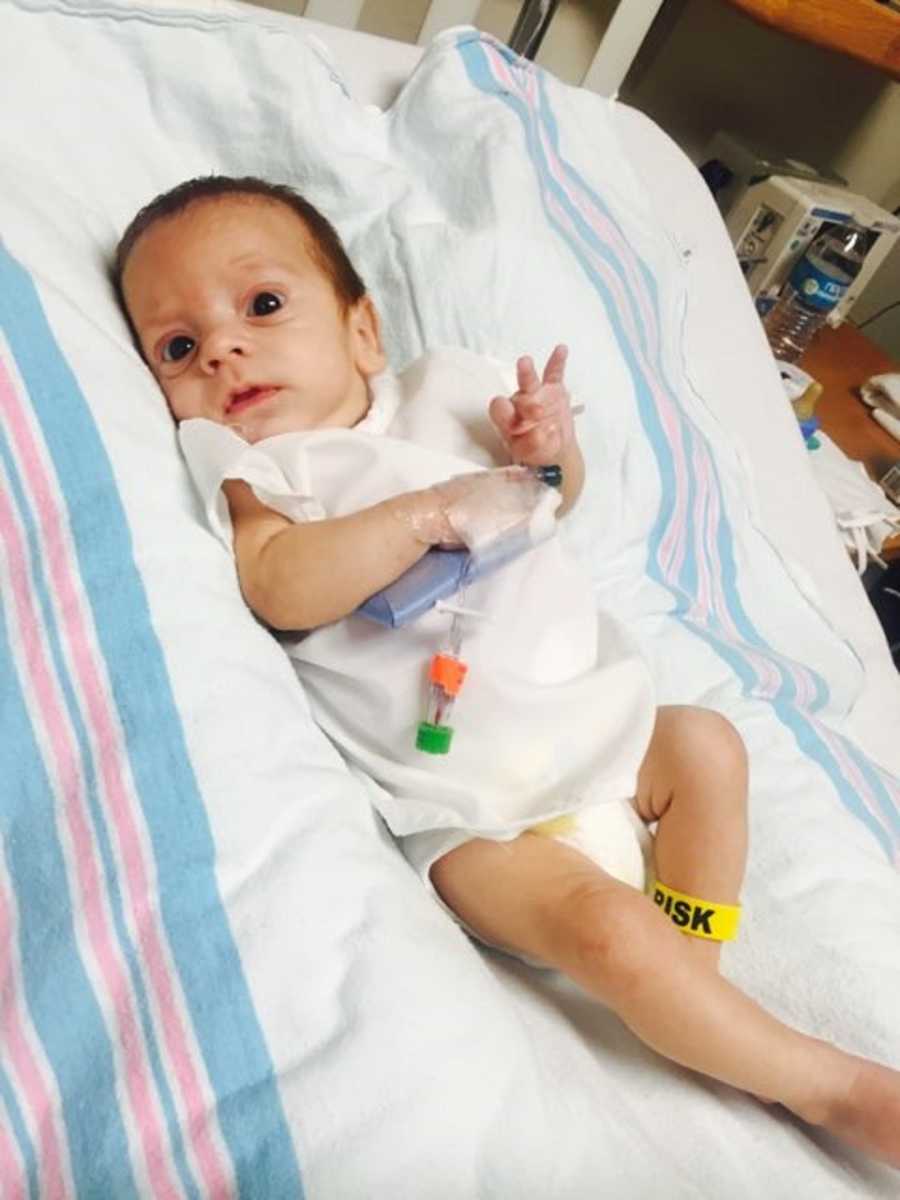 6 week old baby lying in hospital bed with cast on hand and band wrapped around leg that says, "Risk"