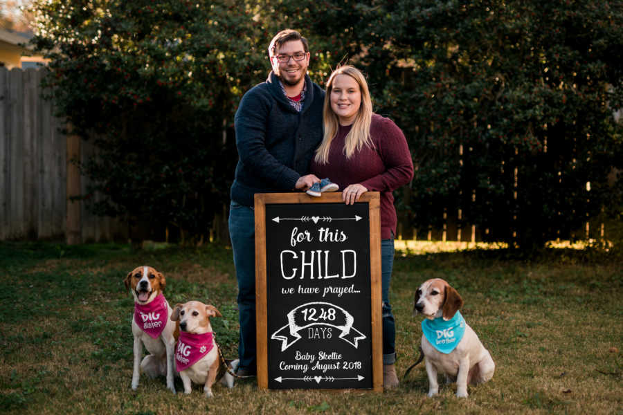 Husband and wife stand behind sign saying, "For this child we have prayed" beside their three dogs