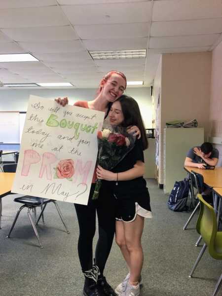 Teen girl holding prom-posal poster in classroom smiles with arm around girlfriend who is holding flowers
