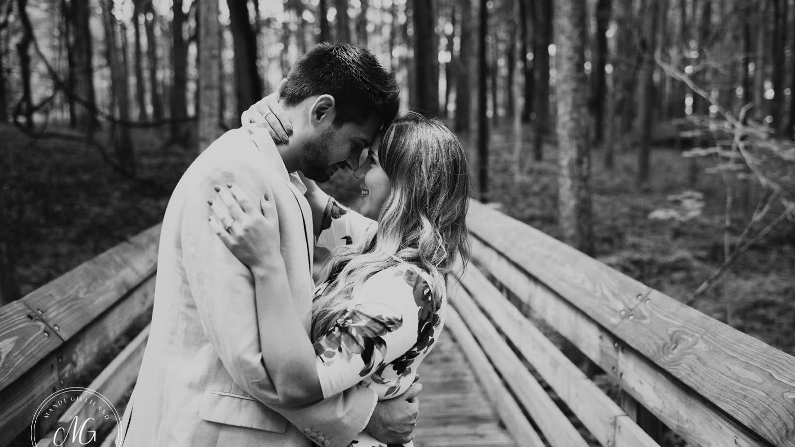 Man and woman stand on wooden bridge in with foreheads together after proposal