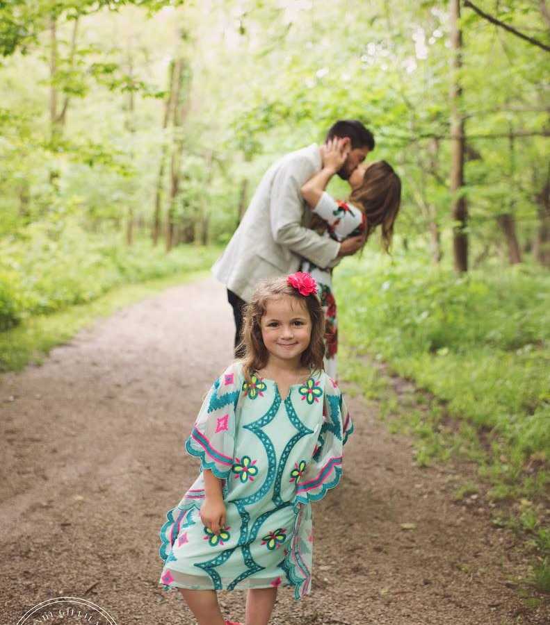 Toddler smiling with hand on hip in front of her mother who was just proposed to and kissing fiancee