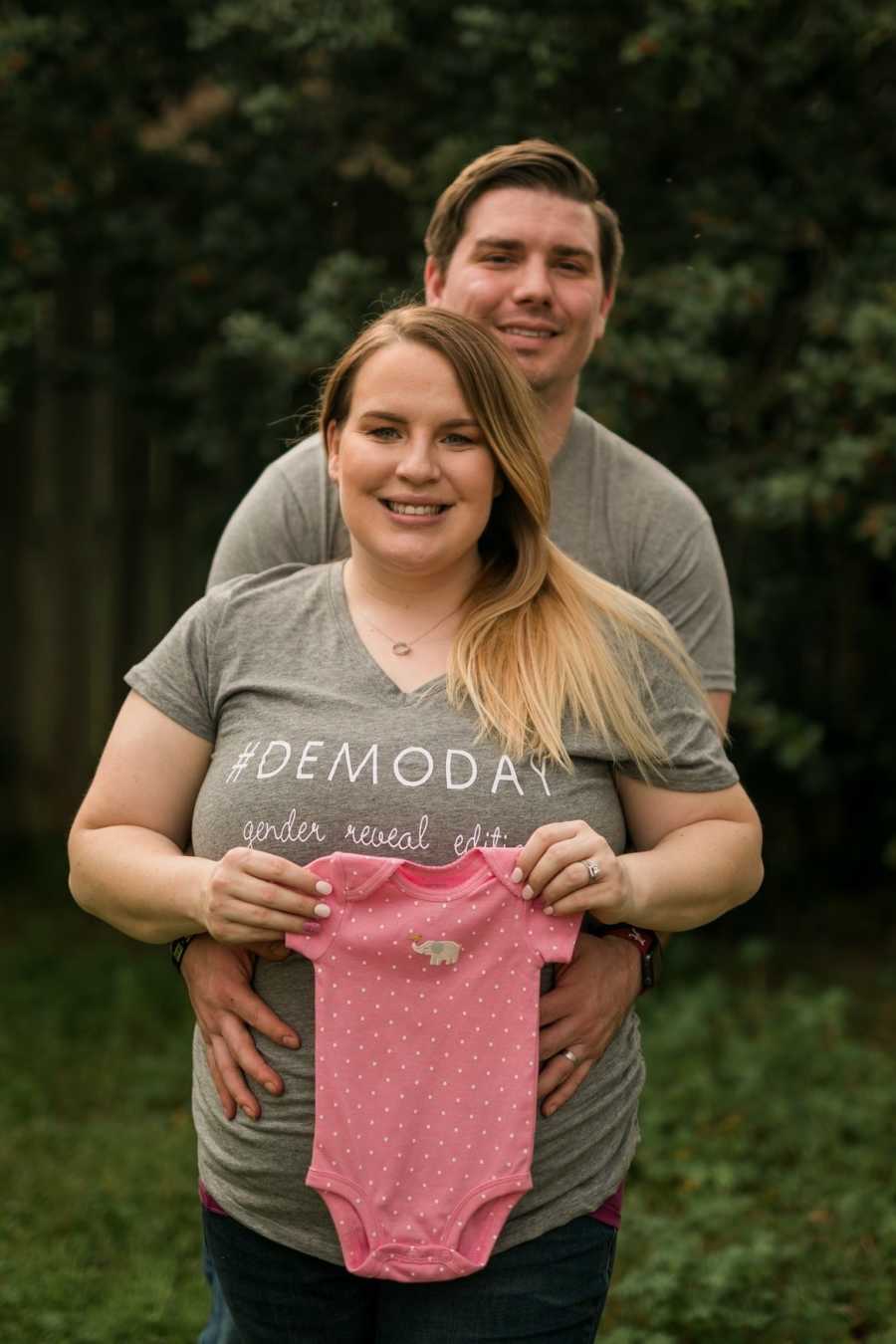 Husband stands with arms around wife who is holding pink onesie for baby who was conceived through IVF