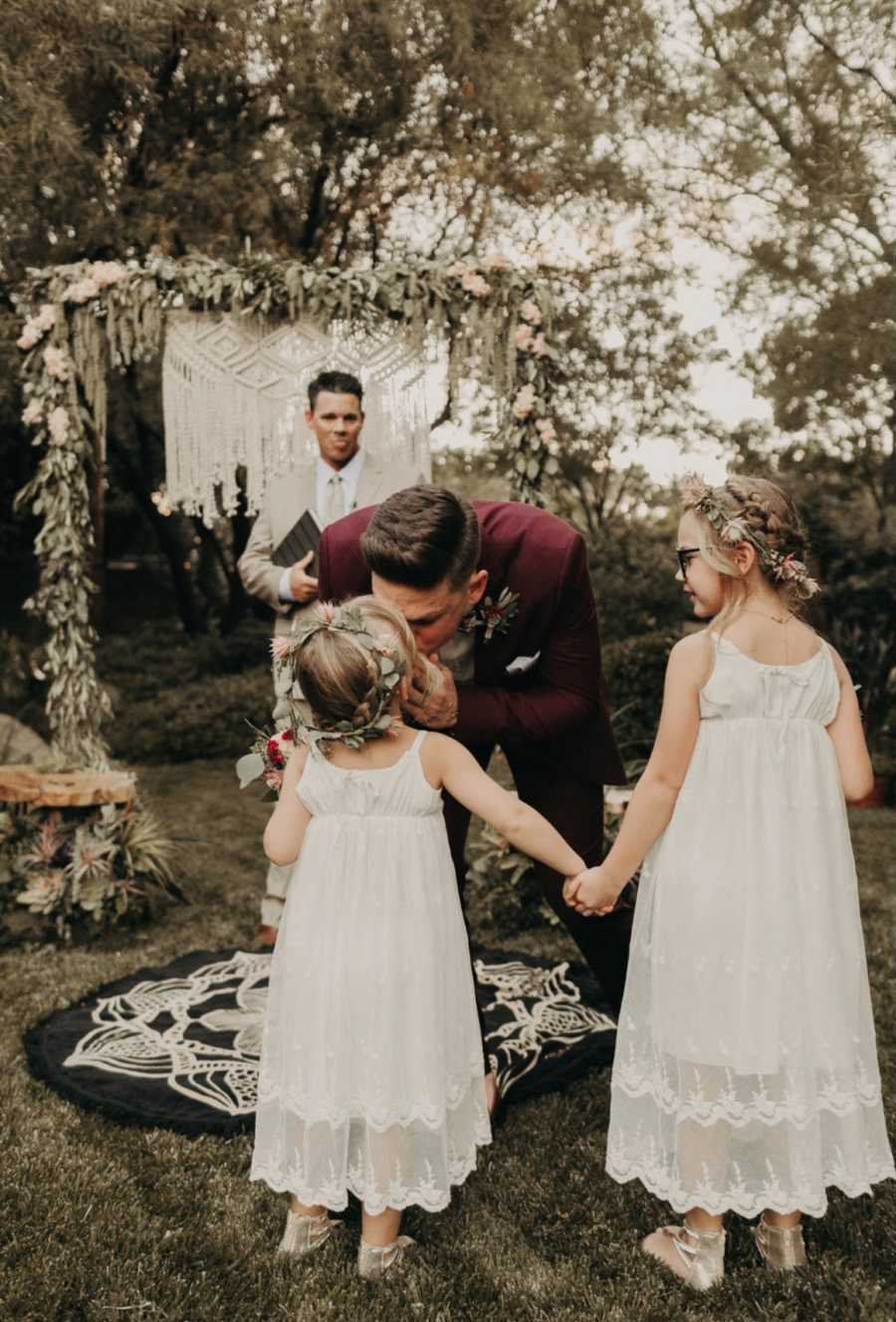 Groom leans over to kiss daughter who is holding hands with half-sister at outdoor wedding