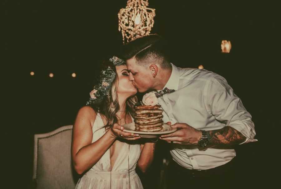 Bride and groom kiss while holding plate with stack of pancakes on it