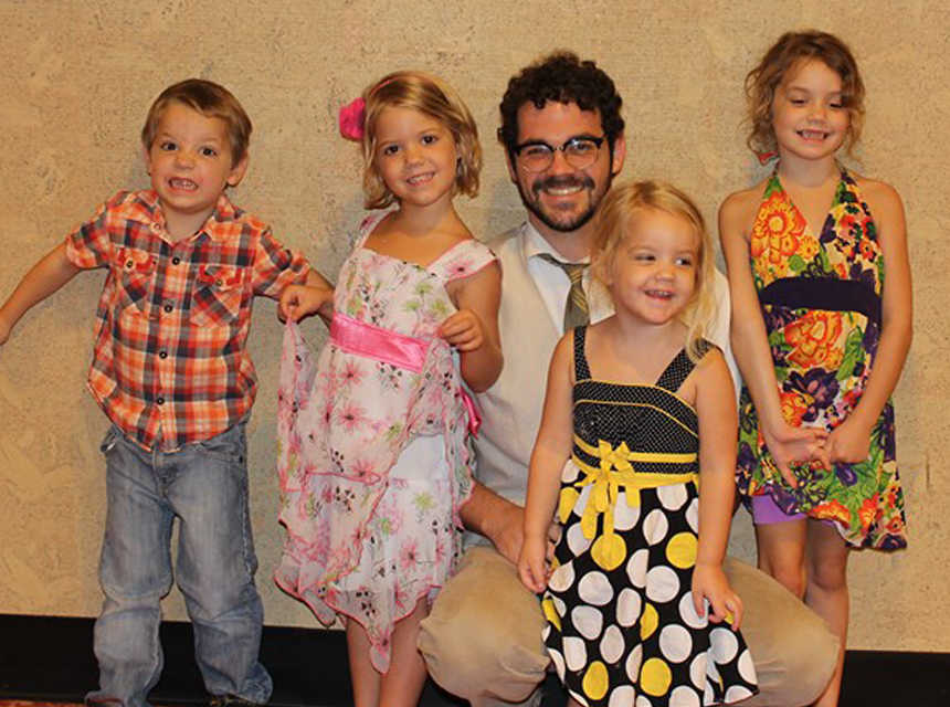 A dad with glasses and a white shirt kneels by his four young children, all of whom are dressed up.