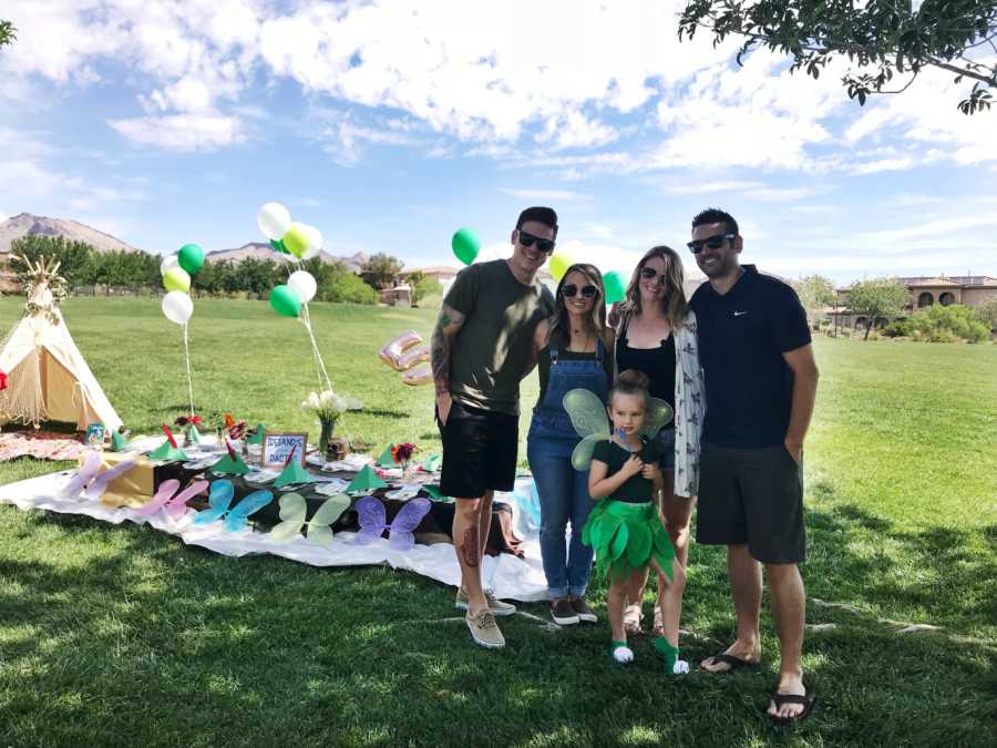 Woman stands outside beside husband, ex-husband and his new wife, while her daughter stands in front of them