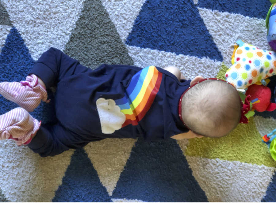 A baby wearing a blue onesie with a rainbow on it lying on a rug with blue and gray triangles