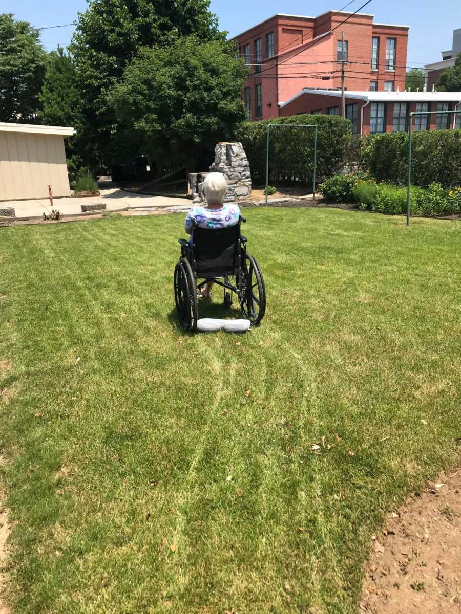 An older woman sits in a wheelchair on bright green grass with her back turned to the camera.