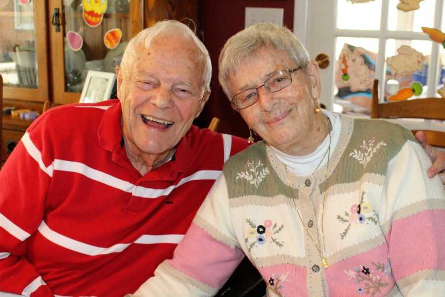 An elderly couple smiling while sitting together in dining room chairs. The man has his arm around his wife.