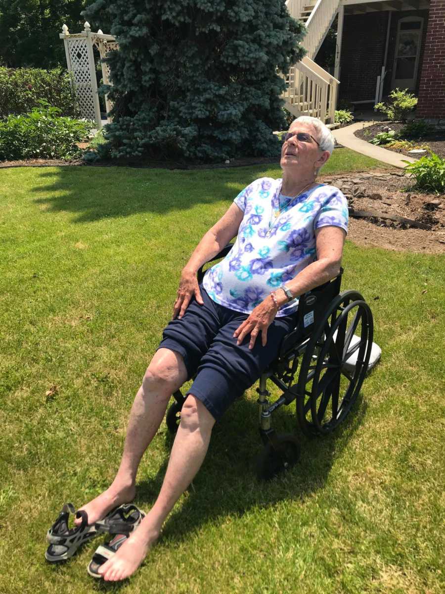 An older woman reclined in a wheelchair in her yard. She wears a floral shirt and rolled-up pants, and her sandals are off.