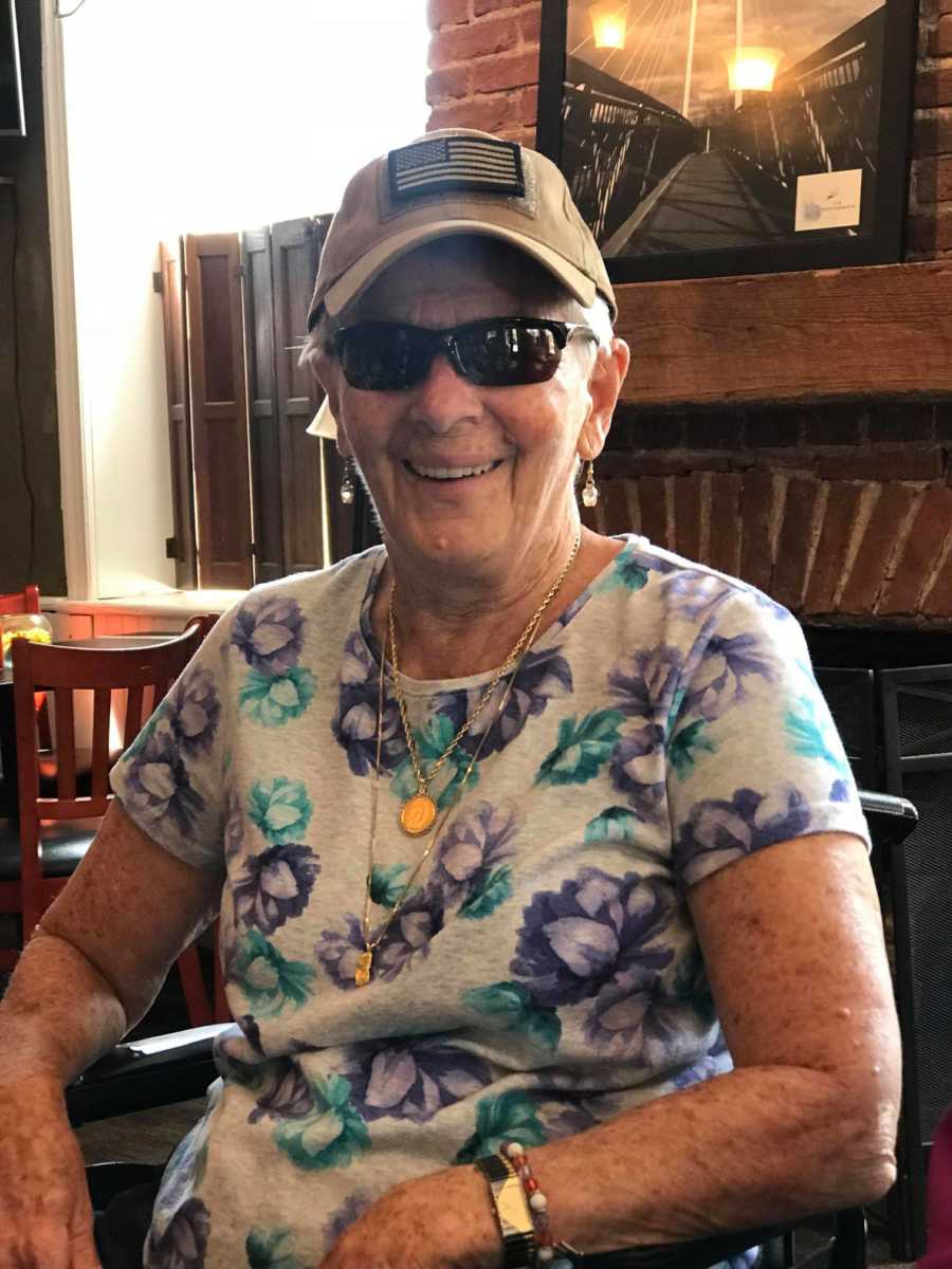 An older woman in a wheelchair wearing a floral shirt, sunglasses, and an American flag ballcap.