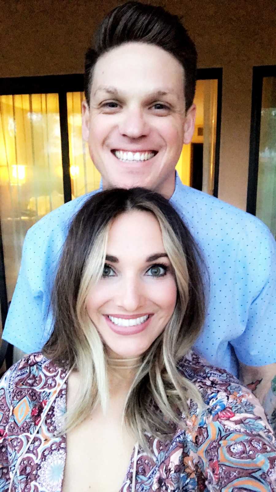 Woman smiles in selfie while her husband stands smiling behind her