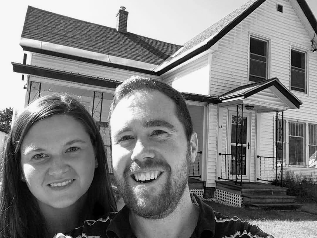 A married man and woman stand in front of a two-story house. The man has a short beard and wears a striped shirt.