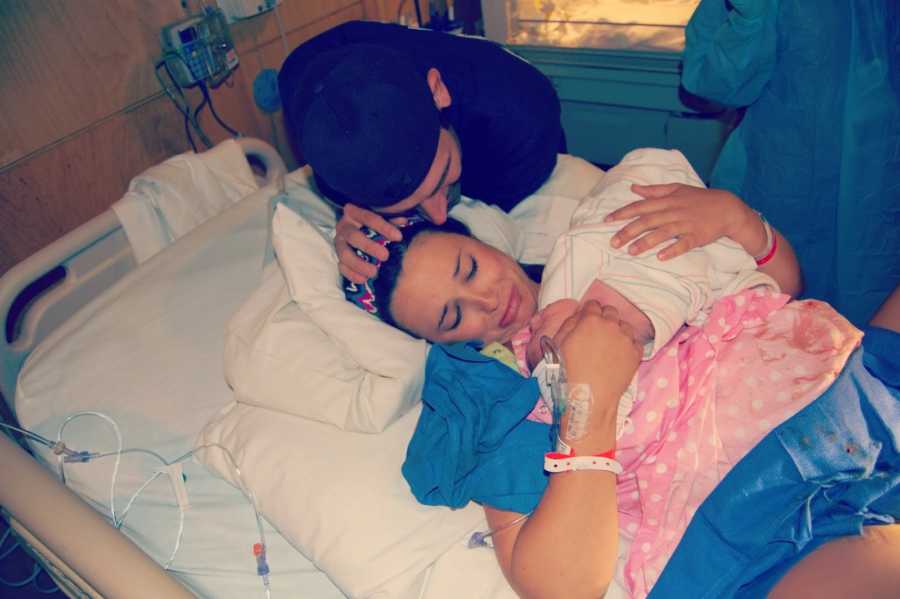 Woman lays in hospital bed crying as she holds newborn while father stands over them