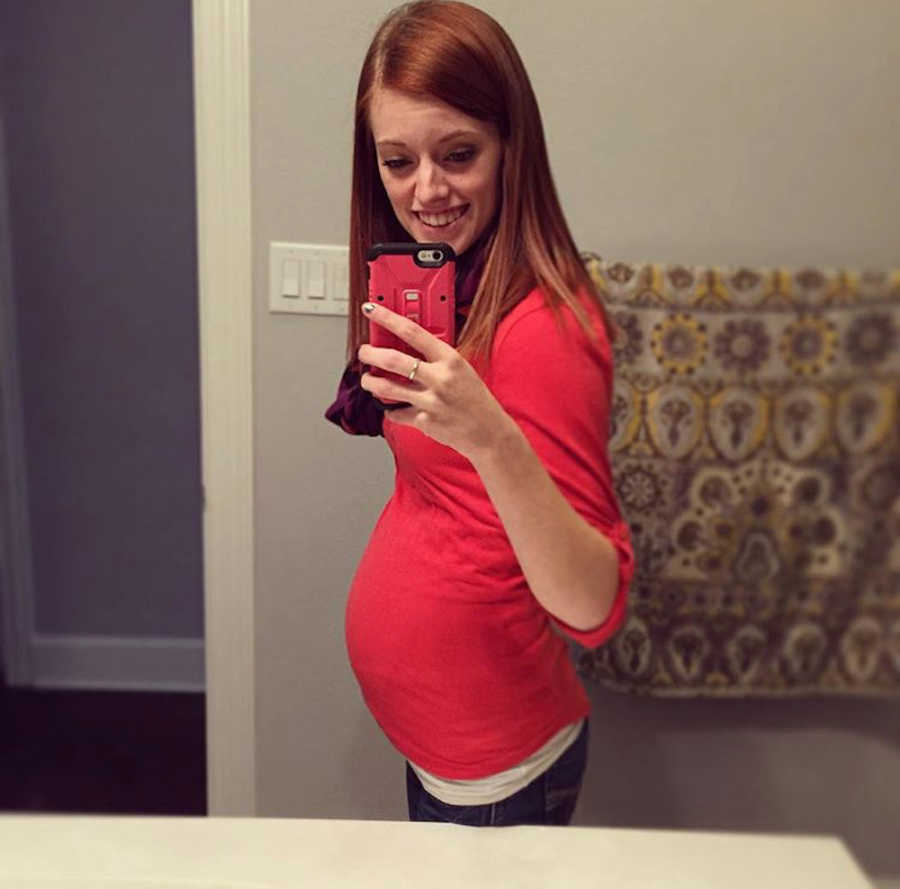 A thin pregnant woman wearing a red shirt takes a photo of herself in the bathroom mirror using her phone.