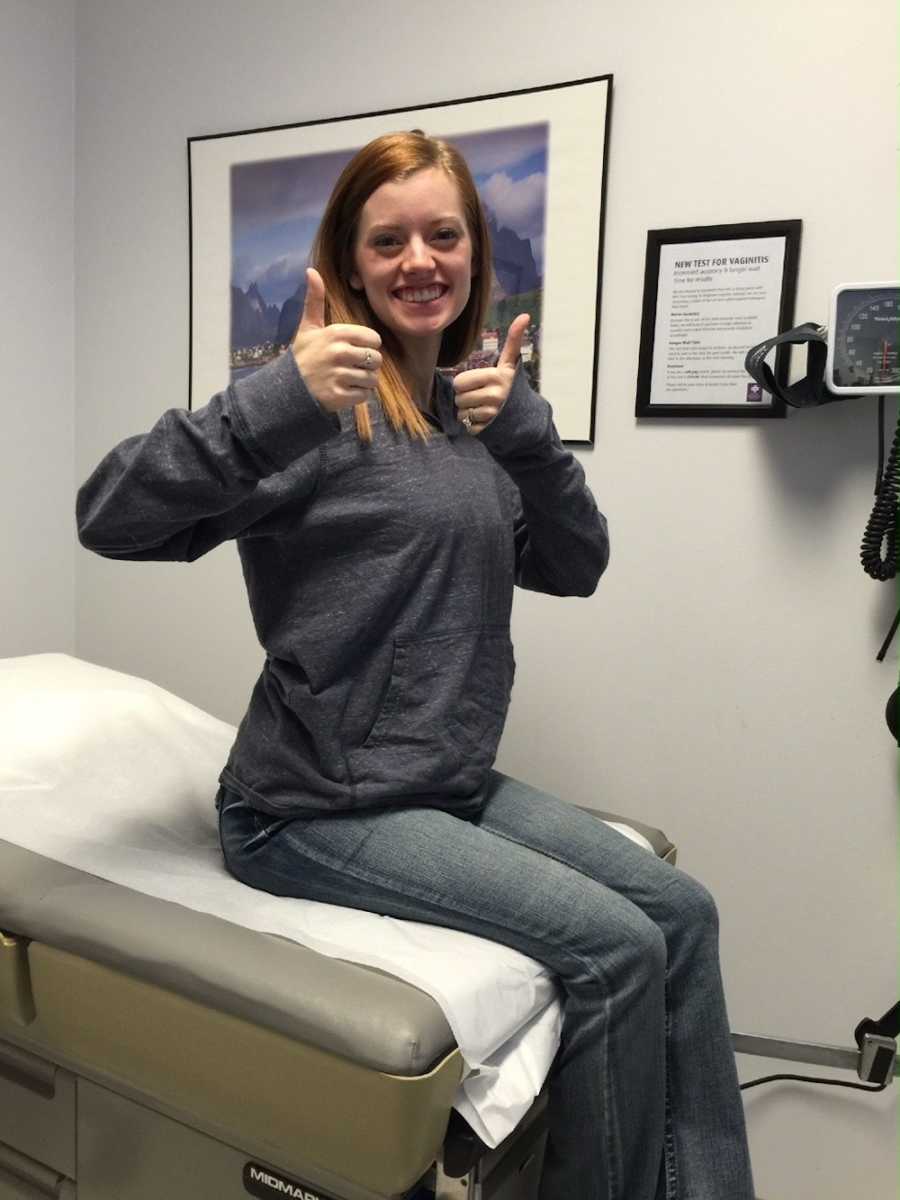 A thin pregnant mom wearing a hoodie and jeans sits on a doctor's table.