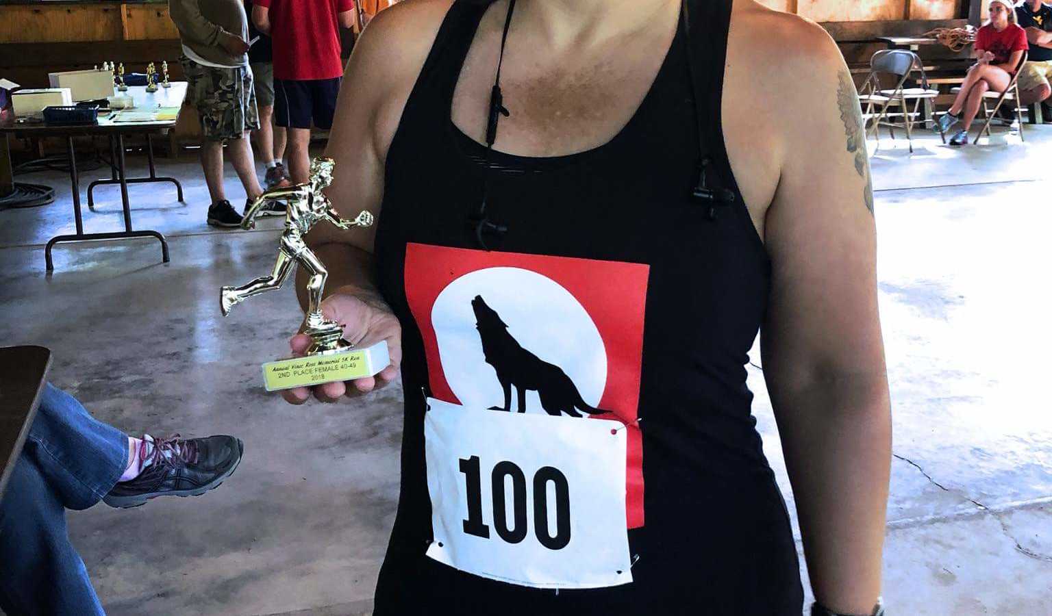 A woman wearing a black tanktop holds a trophy after running a race. On her shirt is a wolf howling.